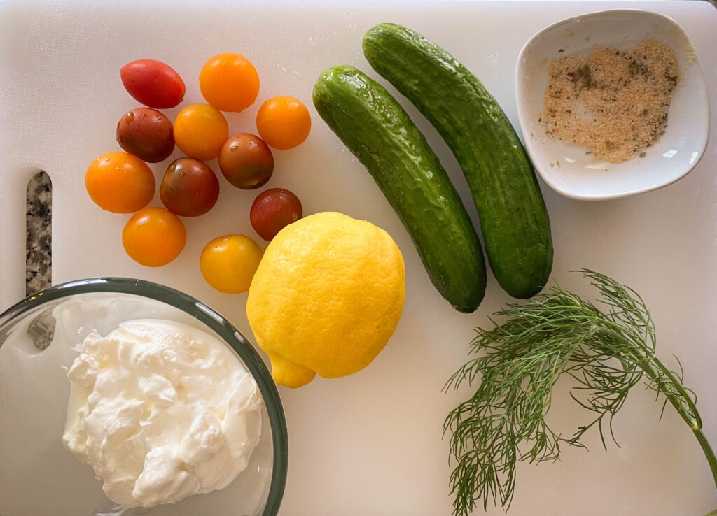 cutting board with sour cream, cherry tomatoes, lemon, cucumbers, fresh dill, and seasonings